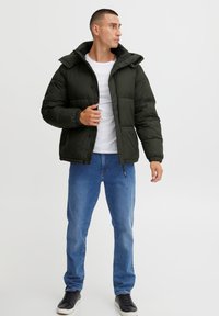 Mørkegrønn overgangsjakke med hette, glidelås og trykknapper; har elastiske mansjetter; kombinert med blå jeans og svarte joggesko.