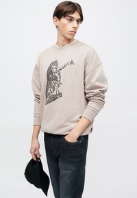 Ανοιχτό μπεζ φούτερ με γραφικό σχέδιο, άνετη εφαρμογή, ριμπ μανσέτες και λαιμόκοψη. Συνδυάζεται με σκούρο τζιν και κρατάει μαύρο καπέλο.