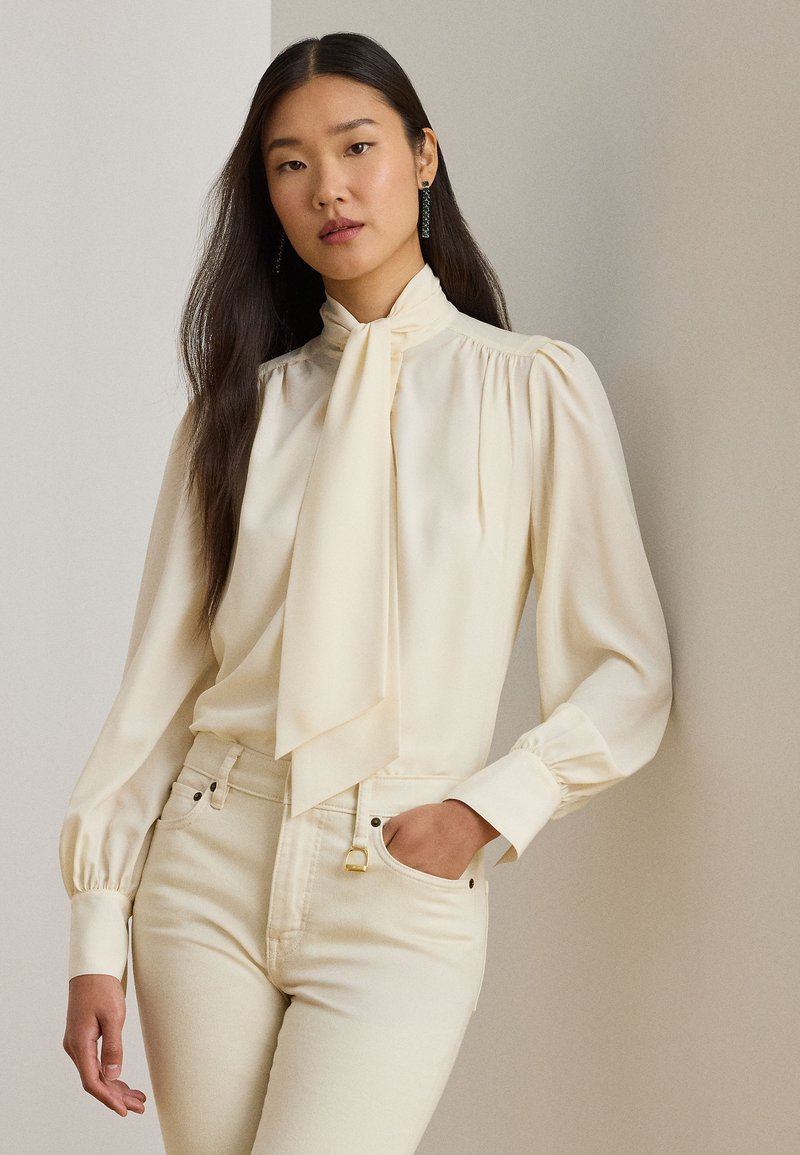 Vrouw met lang donker haar die een crèmekleurige blouse met lange mouwen en een halsstrik draagt en crèmekleurige broek met hoge taille, hand in de zak.