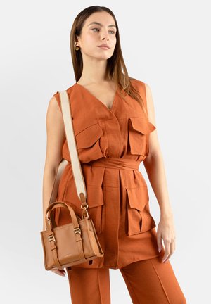 Donna che indossa un gilet utility senza maniche color ruggine e pantaloni, con una borsa a tracolla in pelle color cuoio chiaro con tracolla beige.