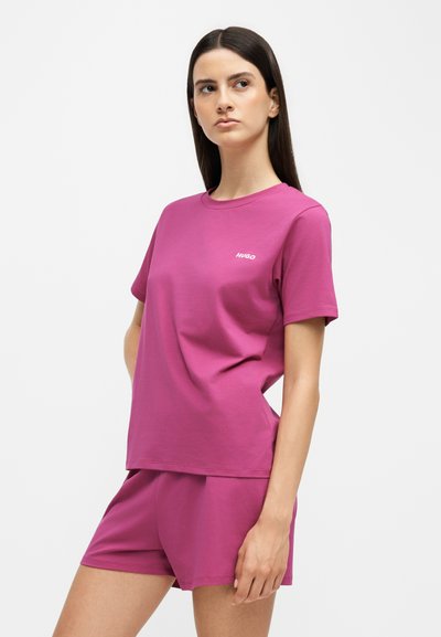 Femme portant un ensemble assorti magenta composé d’un T-shirt à manches courtes et d’un short, avec le logo « Hugo » sur la poitrine, debout devant un fond clair uni.