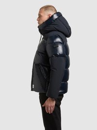 Mornarsko modra puhovka z bleščečim zaključkom, velikim kapuco in stranskimi žepi z zadrgo. Dizajn ima kviltano teksturo in sega do kolen.