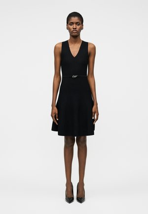 Kobieta stojąca w czarnej, bezrękawowej sukience z dekoltem w serek, z cienkim paskiem i czarnymi, spiczastymi szpilkami na jednolitym białym tle.