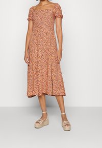 Robe midi florale avec des manches bouffantes courtes, décolleté carré, base orange et motifs de petites fleurs multicolores. Associée à des sandales compensées.