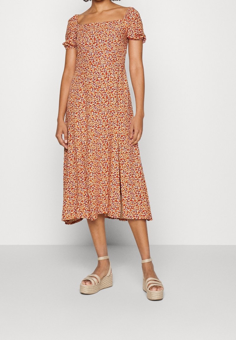 Robe midi florale avec des manches bouffantes courtes, décolleté carré, base orange et motifs de petites fleurs multicolores. Associée à des sandales compensées.