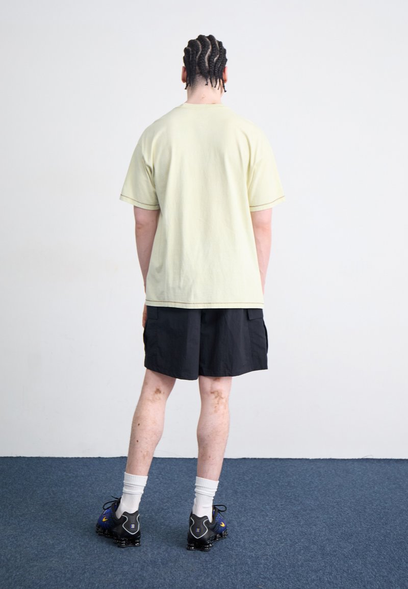 T-shirt en coton jaune clair, shorts cargo noirs, chaussettes blanches et chaussures de sport noires avec des accents bleus. Coiffure tressée.