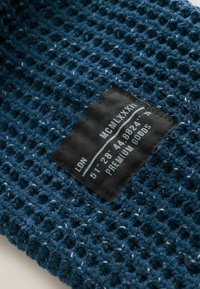 Tessuto blu tessuto con un motivo a nido d'ape, dotato di un'etichetta nera con testo bianco che fornisce istruzioni per la cura e informazioni sul marchio.