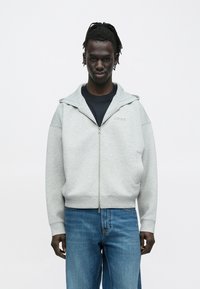 Γκρι φούτερ με φερμουάρ και κουκούλα, με ομαλή υφή και μανίκια σε αντίθεση. Φορεμένο πάνω από μαύρο πουκάμισο, σε συνδυασμό με ανοιχτόχρωμο μπλε τζιν.