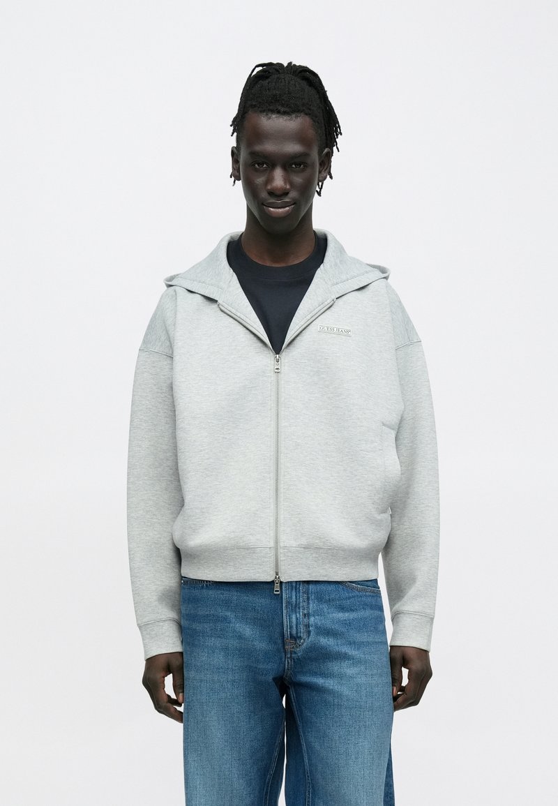 Γκρι φούτερ με φερμουάρ και κουκούλα, με ομαλή υφή και μανίκια σε αντίθεση. Φορεμένο πάνω από μαύρο πουκάμισο, σε συνδυασμό με ανοιχτόχρωμο μπλε τζιν.