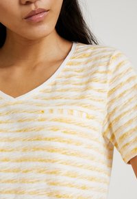 Camiseta de algodón de manga corta con escote en V, con una base blanca decorada con rayas amarillas onduladas y un pequeño bolsillo horizontal en el pecho.