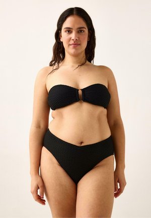 Kvinde med langt, mørkt hår iført en tekstureret sort bikini top uden stropper og bikinitrusser med høj talje, stående mod en ensfarvet baggrund.