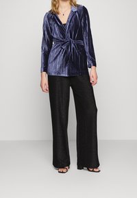 Navy fluwelen wikkeltrui met lange mouwen en een strikaccent, gecombineerd met zwarte glitterwijde pijpen en heeled sandalen.