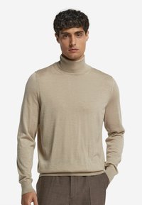 Beżowy sweter z golfem, wykonany z miękkiego materiału, z długimi rękawami, dopasowanym krojem i prążkowanym kołnierzem. Sweter zestawiony z brązowymi spodniami.