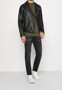 Giacca da moto nera in pelle con zip argentate, maglione verde oliva, jeans scuri e sneakers bianche. Texture liscia, design attillato.