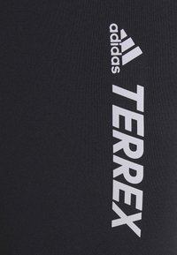 Logo Adidas Terrex en blanc sur tissu noir texturé, avec l'emblème à trois bandes de la marque au-dessus du texte.