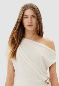 Top bianco scivolato sulle spalle con design drappeggiato, caratterizzato da un accento dorato in vita e una texture morbida del tessuto. Forma liscia ed elegante.