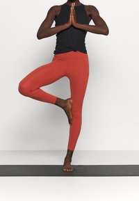 Mustat ribbitoppi ja ruosteenväriset trikoot. Trikoot ovat vartalonmyötäiset, sileäpintaiset ja korkeavyötäröiset. Asento on puun tasapaino.