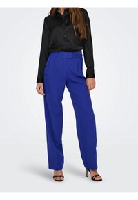 Chemise en satin noir à boutons avec manches longues associée à un pantalon taille haute à jambes larges de couleur bleu vif, portée avec des talons noirs à lanières.