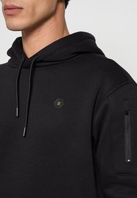 Zwarte hoodie met een capuchon met trekkoord, een zeshoekig logo op de borst en een zak met rits op de mouw. Heeft een getextureerde stof.