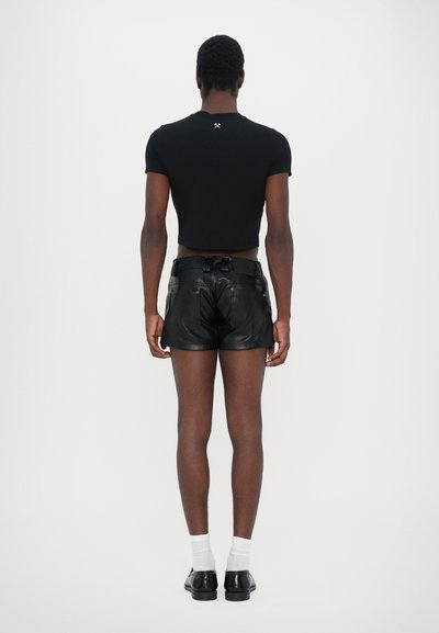 Tricou scurt negru cu un mic logo pe spate, asortat cu pantaloni scurți din piele neagră, prevăzuți cu buzunare și bucle pentru curea. Șosete albe și pantofi negri.