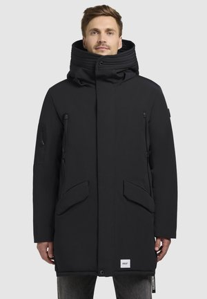 Parka negra aislante con un cuello alto, dos bolsillos frontales, un bolsillo con cremallera en el pecho y un parche de logotipo cerca del dobladillo. Textura lisa y mate.