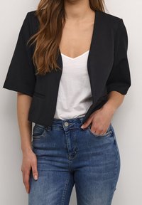 Svart kort kavaj med korta ärmar, ihop med en vit V-ringad tröja och blå jeans. Slät tyg med minimal detaljering.