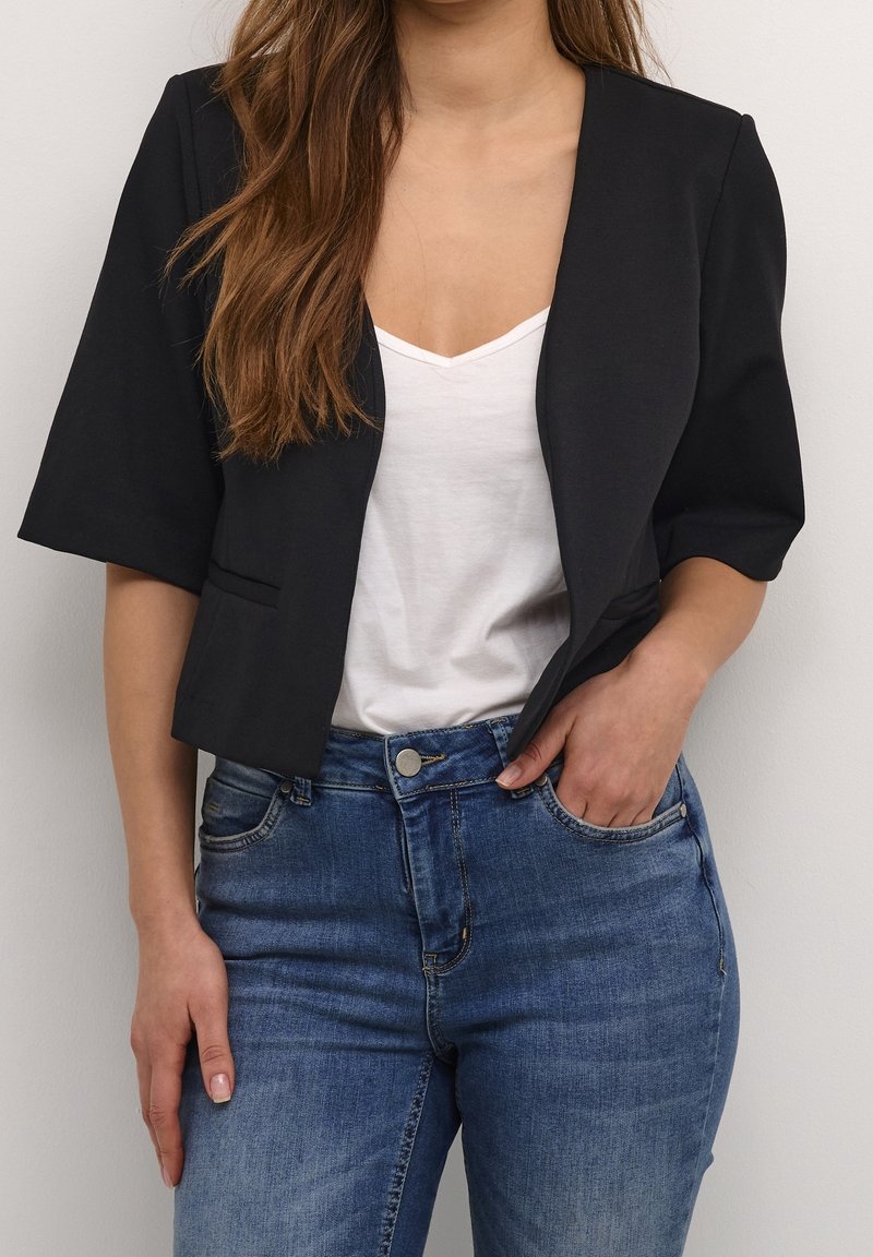 Svart kort kavaj med korta ärmar, ihop med en vit V-ringad tröja och blå jeans. Slät tyg med minimal detaljering.