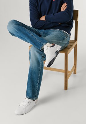 Personne portant un jean bleu, des baskets blanches et un sweat-shirt bleu marine assise en tailleur sur une chaise en bois devant un fond uni.