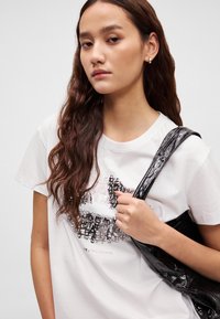 Hvit t-skjorte med grafisk trykk som viser svart tekst og abstrakte mønstre. Modellen bærer en blank svart veske med teksturerte stropper.