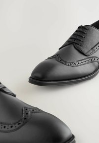 Czarne skórzane buty eleganckie z gładką cholewką, ozdobnym przeszyciem w stylu brogue, spiczastym przodem i okrągłymi sznurówkami z teksturowaną naszą. 