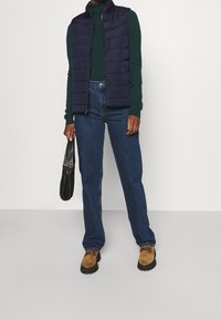 Persoon draagt een marineblauwe doorgestikte bodywarmer over een groen shirt met lange mouwen, donkerblauwe spijkerbroek, beige loafers en houdt een kleine zwarte handtas vast.