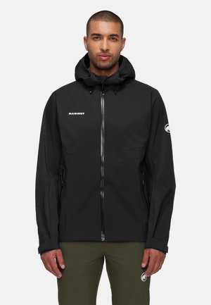 Mann trägt schwarze Mammut-Kapuzenjacke mit Reißverschluss vorne und olivgrüne Hose vor einfachem Hintergrund.