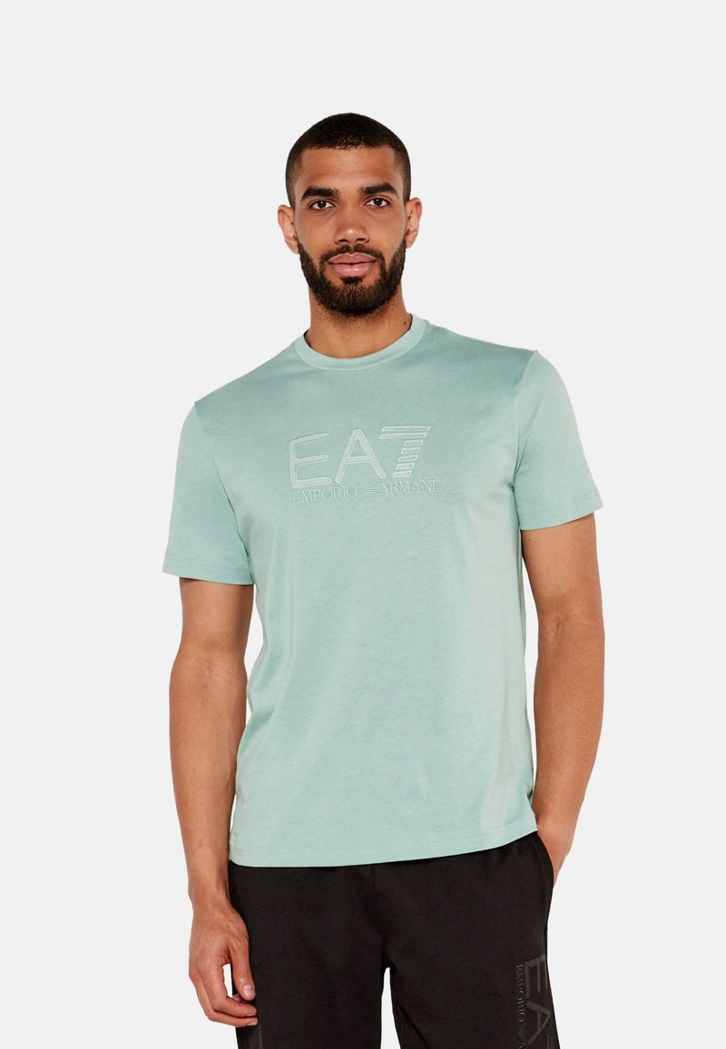 Camiseta de cuello redondo verde claro hecha de algodón suave, con un logo EA7 en relieve en el pecho y mangas cortas. Llevada con pantalones negros.