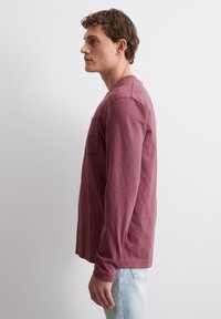 Langärmliges, kastanienrotes Hemd aus strukturiertem Stoff mit Rundhalsausschnitt und einer Brusttasche, kombiniert mit hellblauen Jeans.