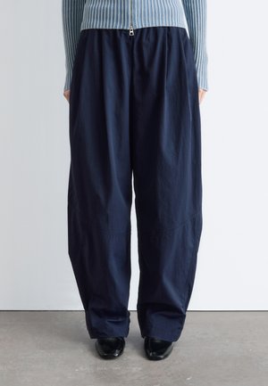 Osoba nosząca luźne, ciemnogranatowe spodnie zestawione z czarnymi butami i jasnoniebieskim, prążkowanym swetrem na zamek, stojąca na szarej podłodze.