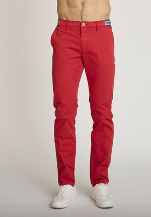Homme portant un pantalon slim rouge avec une ceinture à motif bleu et blanc, associé à des baskets blanches, debout devant un fond uni.