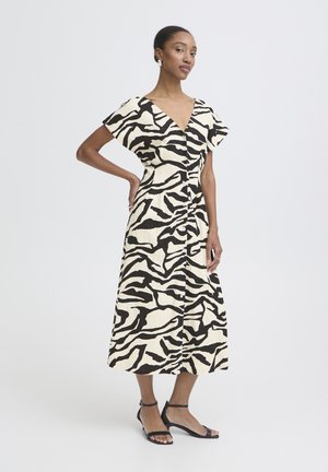 Femme grande portant une robe midi à imprimé zèbre noir et blanc et des talons noirs à brides, posant avec une main sur la hanche.