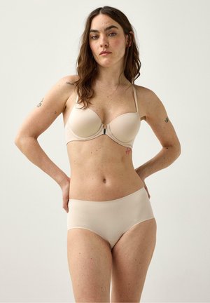 Kvinde med langt brunt hår og tatoveringer iført beige bh og trusse, stående med hænderne bag ryggen mod en lys baggrund.