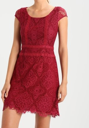 Femme portant une robe en dentelle bordeaux près du corps avec des manches courtes, des motifs floraux et un ourlet festonné, debout devant un fond uni.