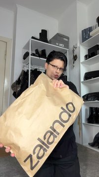 Saco de compras da Zalando em papel castanho segurado por uma pessoa, apresentando um logótipo preto em destaque. O fundo inclui prateleiras com sapatos e acessórios.