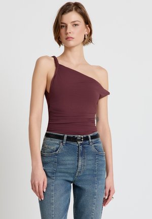 Giovane donna con capelli corti che indossa un top bordeaux monospalla senza maniche e jeans blu a vita alta con cintura nera.