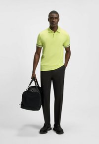 Limegrön pikétröja med svartvita randiga detaljer, svarta byxor och en svart duffelväska; modellen står självsäkert och bär svarta sneakers.