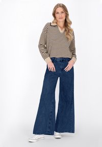 Sweter w paski w beżowo-czarnym kolorze z dekoltem w serek, zestawiony z szerokimi niebieskimi dżinsami. Model nosi białe sneakersy. Prosta, casualowa stylizacja.