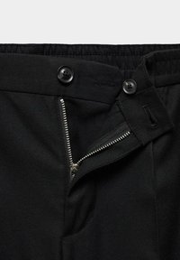 Pantalon noir avec une fermeture éclair argentée non zippée et deux boutons noirs sur la ceinture, dont un attaché et un détaché.