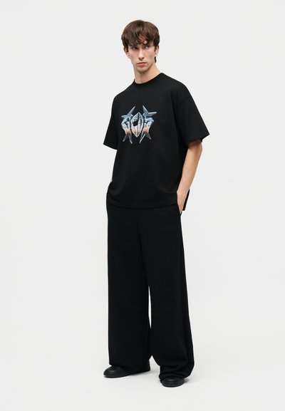 Νεαρός άνδρας φοράει φαρδιά μαύρη μπλούζα με σχέδιο και μαύρο παντελόνι με φαρδιά μπατζάκια, στέκεται με τα χέρια στις τσέπες μπροστά σε ουδέτερο φόντο.