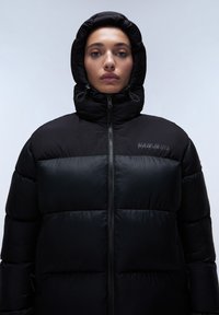 Giacca a vento nera con collo alto e cappuccio. Design trapuntato, zip frontale e dettaglio del logo sulla parte superiore sinistra. Texture liscia e lucida.