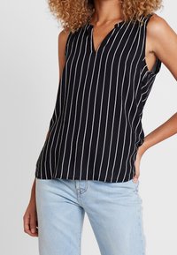 Ärmlös blus i svart och vitt randigt mönster med V-ringning och mjukt tyg. Har en avslappnad passform och subtila sidoklyftor vid fållen.