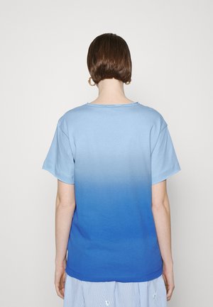 Person mit kurzen braunen Haaren, die goldene Creolen und ein blaues Ombre-Kurzarmhemd trägt, von hinten vor einem schlichten Hintergrund gesehen.