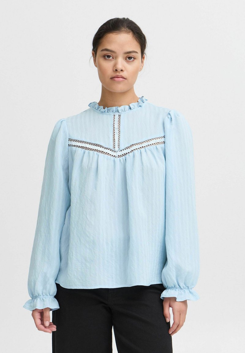 Blusa azzurra chiara a maniche lunghe con scollatura e polsini a volante. Il tessuto testurizzato presenta dettagli in pizzo sul petto e sulle spalle.
