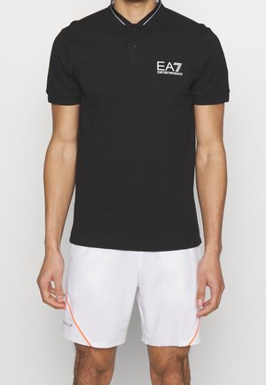 Mann trägt schwarzes EA7 Emporio Armani Poloshirt und weiße Shorts mit orangen Details, steht vor einem einfarbigen Hintergrund.